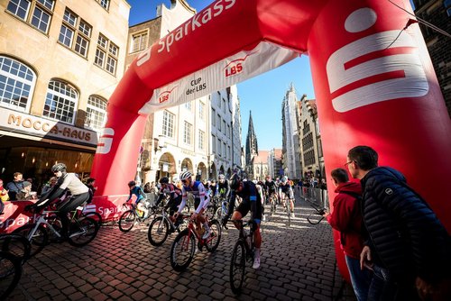 Radrennfahrer starten unter einem roten Torbogen auf dem Prizipalmarkt in Münster vor Zuschauern.