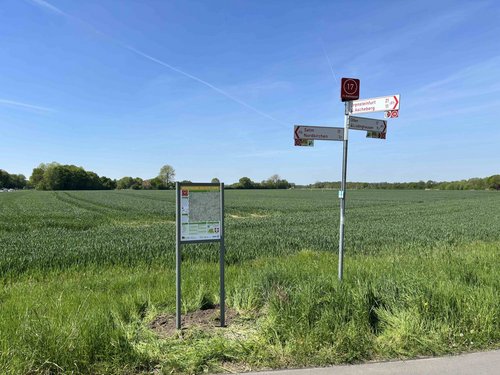 Am Rand eines asphaltierten Weges stehen eine Übersichtstafel mit Radwegenetzkarte und ein Wegweiser mit mehreren rot-weißen Schildern. Im Hintergrund ist eine grüne, weite Feldlandschaft unter blauem Himmel zu sehen.