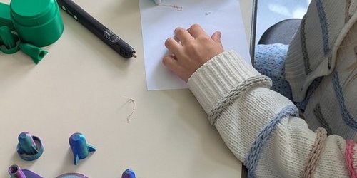 Man sieht die Hände eines Kindes, das einen großen Stift hält. Aus dem Stift kommt ein festes Material, das strichförmig auf einem Blatt Papier liegt.