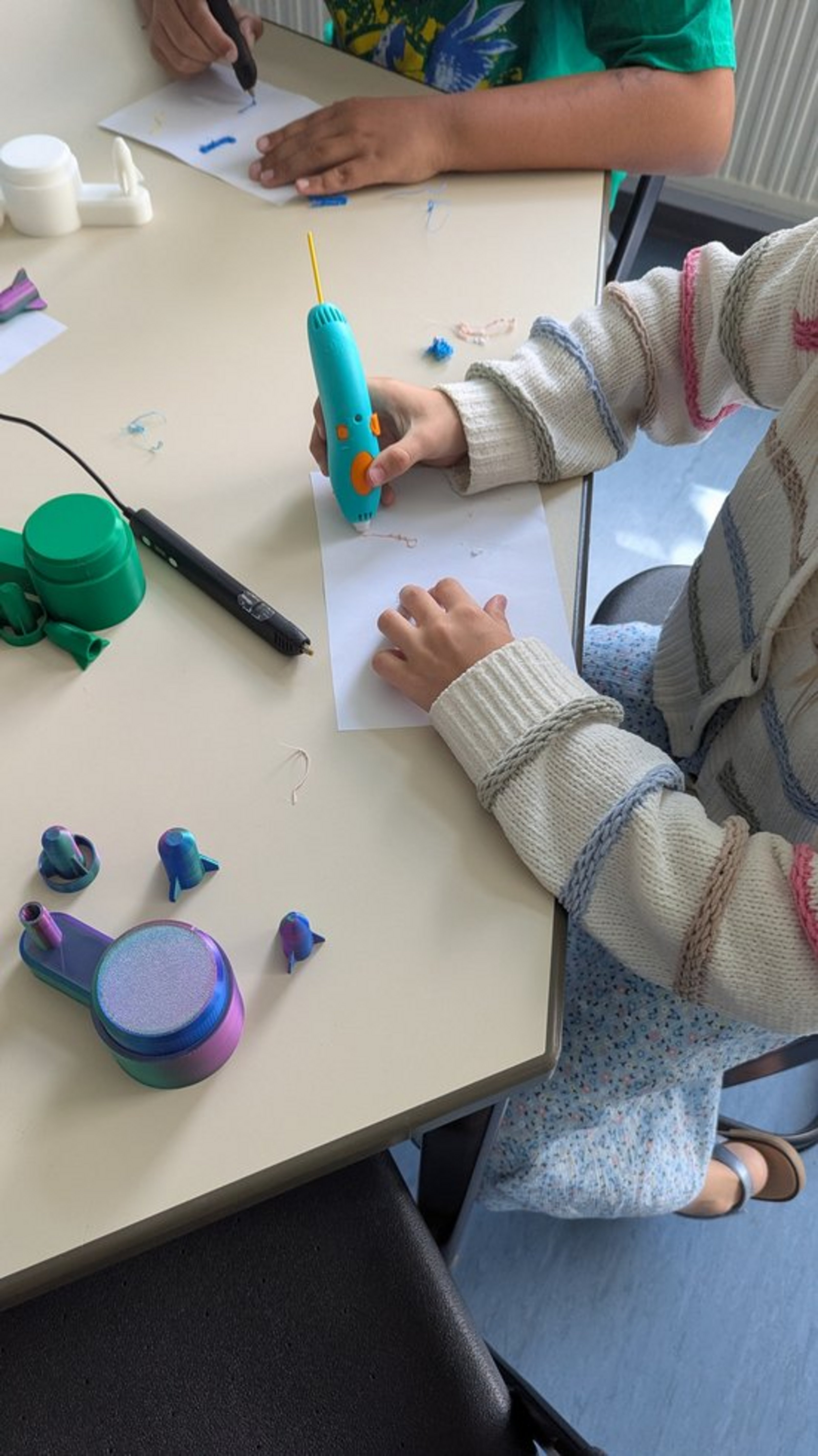 Man sieht die Hände eines Kindes, das einen großen Stift hält. Aus dem Stift kommt ein festes Material, das strichförmig auf einem Blatt Papier liegt.