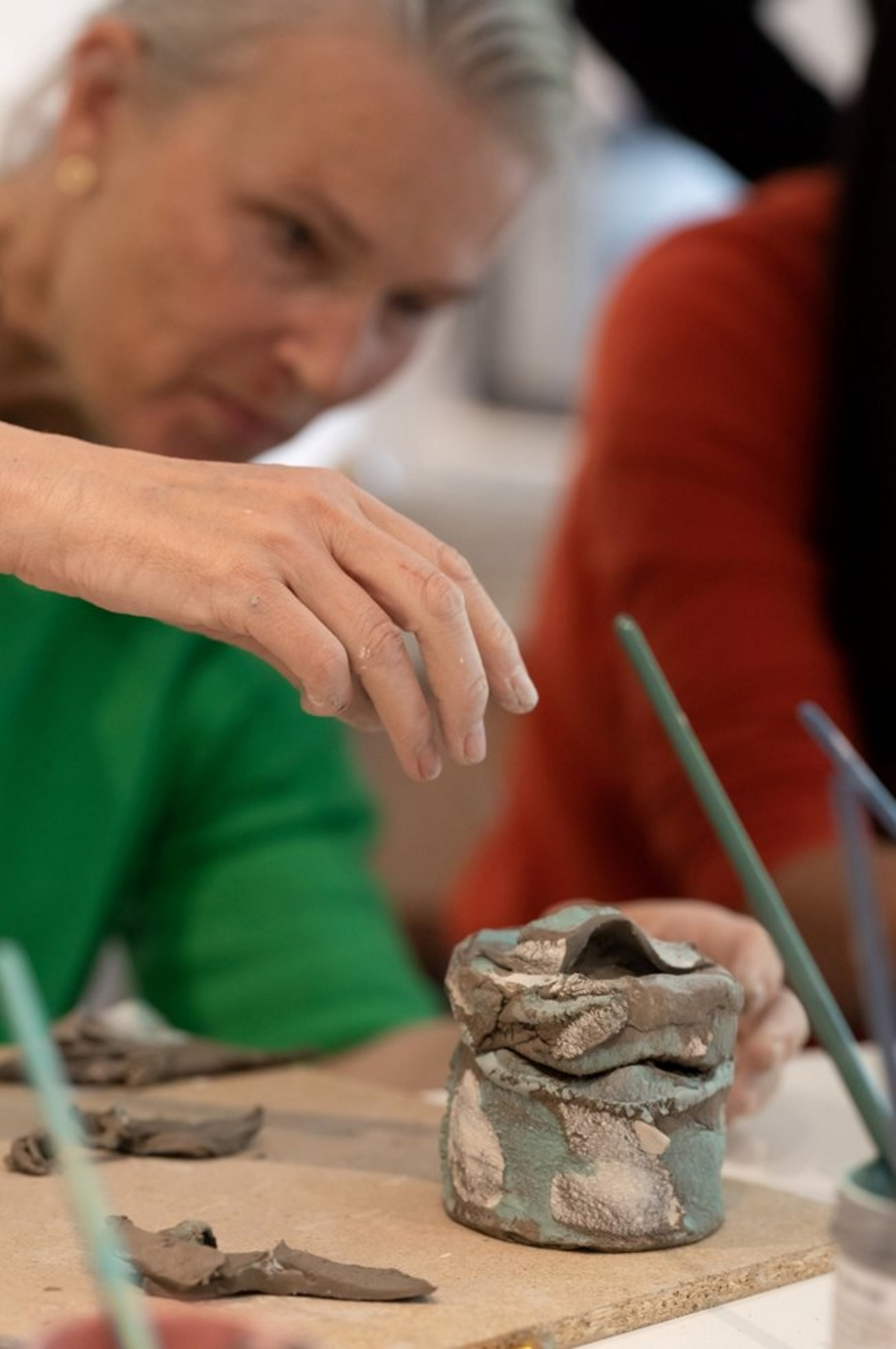 Eine Frau in grünem Oberteil hat ein Gefäß aus frischem Ton vor sich stehen. Auf dem Tisch vor ihr befindet sich außerdem ein Holzbrett, auf dem weitere Tonstücke liegen.