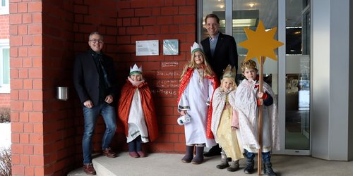 Zwei Männer und vier Kinder in Sternsinger-Gewändern und mit einem Stern  stehen vor dem Eingang eines Gebäudes.