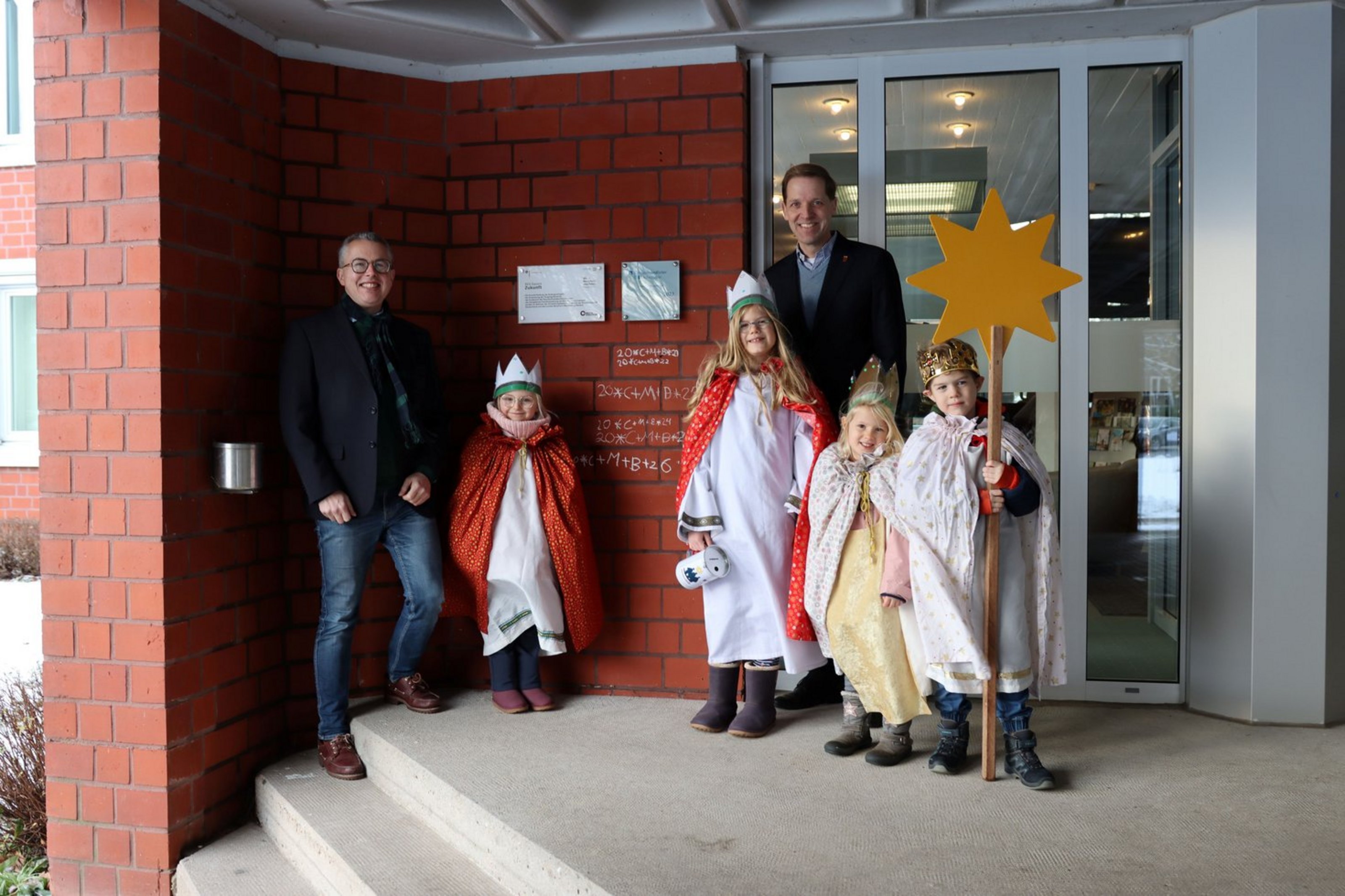 Zwei Männer und vier Kinder in Sternsinger-Gewändern und mit einem Stern  stehen vor dem Eingang eines Gebäudes.