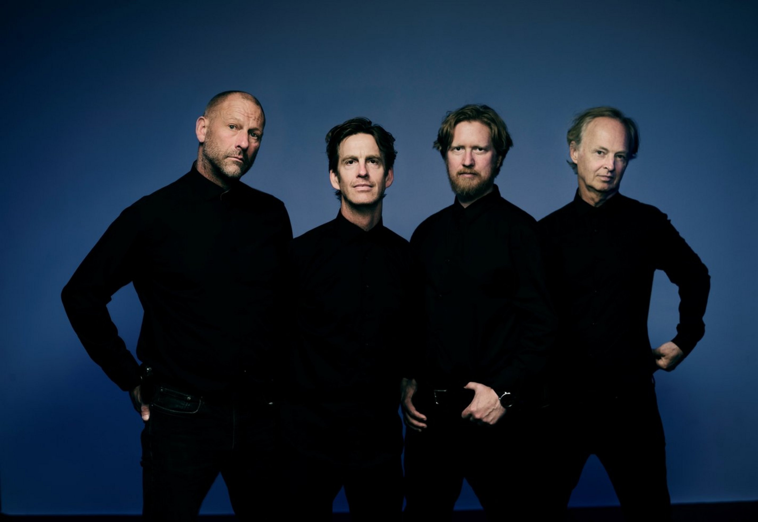 Vier Männer in dunkler Kleidung stehen vor einem blauen Hintergrund. Die Hände haben sie zum Teil auf die Hüften  gestützt.