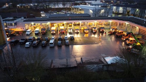 Luftbild des Bauhofgeländes in Dülmen-Buldern in der Dämmerung. Auf dem Platz stehen ordentlich zahlreiche Fahrzeuge, bei denen größtenteils die Beleuchtung eingeschaltet ist. 