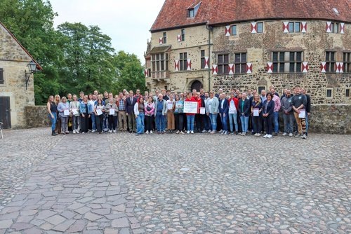 Eine Gruppe von Menshcen steht vor einer Burg auf einem Vorplatz. Einige Personen halten Schilder in den Händen.