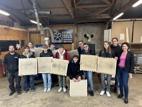 Eine Gruppe von dreizehn Personen steht in einer Werkstatt. Die meisten der Personen sind Jugendliche, begleitet von mehreren Erwachsenen. Sie halten quadratische Holzplatten mit einem Raster aus kleinen, quadratischen Vertiefungen. Im Hintergrund sind Werkzeuge, Maschinen und ein Werbeschild mit dem Logo und Namen „Kawentsmann“ sichtbar.