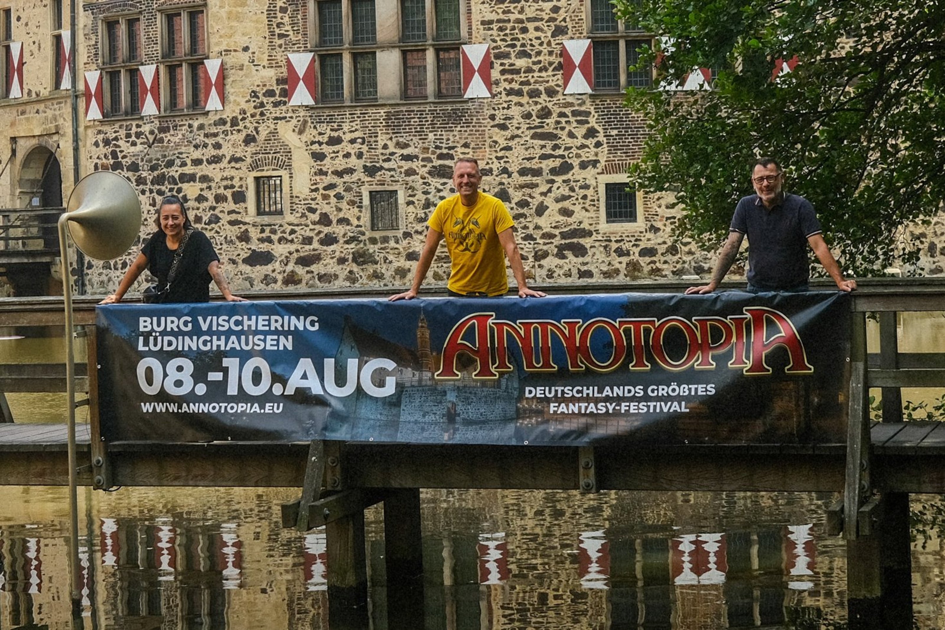 Drei Personen stehen auf einer Holzbrücke vor einer historischen Burg und halten ein Banner für das Fantasyfestival „Annotopia“ in Lüdinghausen vom 8. bis 10. August.