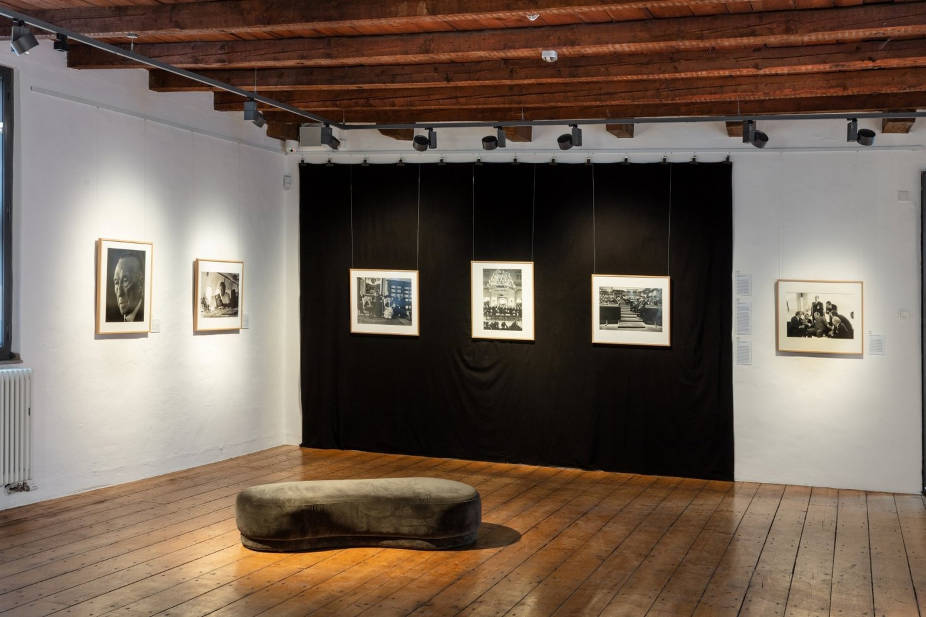 In einem Raum mit Holzboden und zwei weißen sowie einer schwarzen Wand hängen sechs Fotografien gerahmt an den Wänden.
