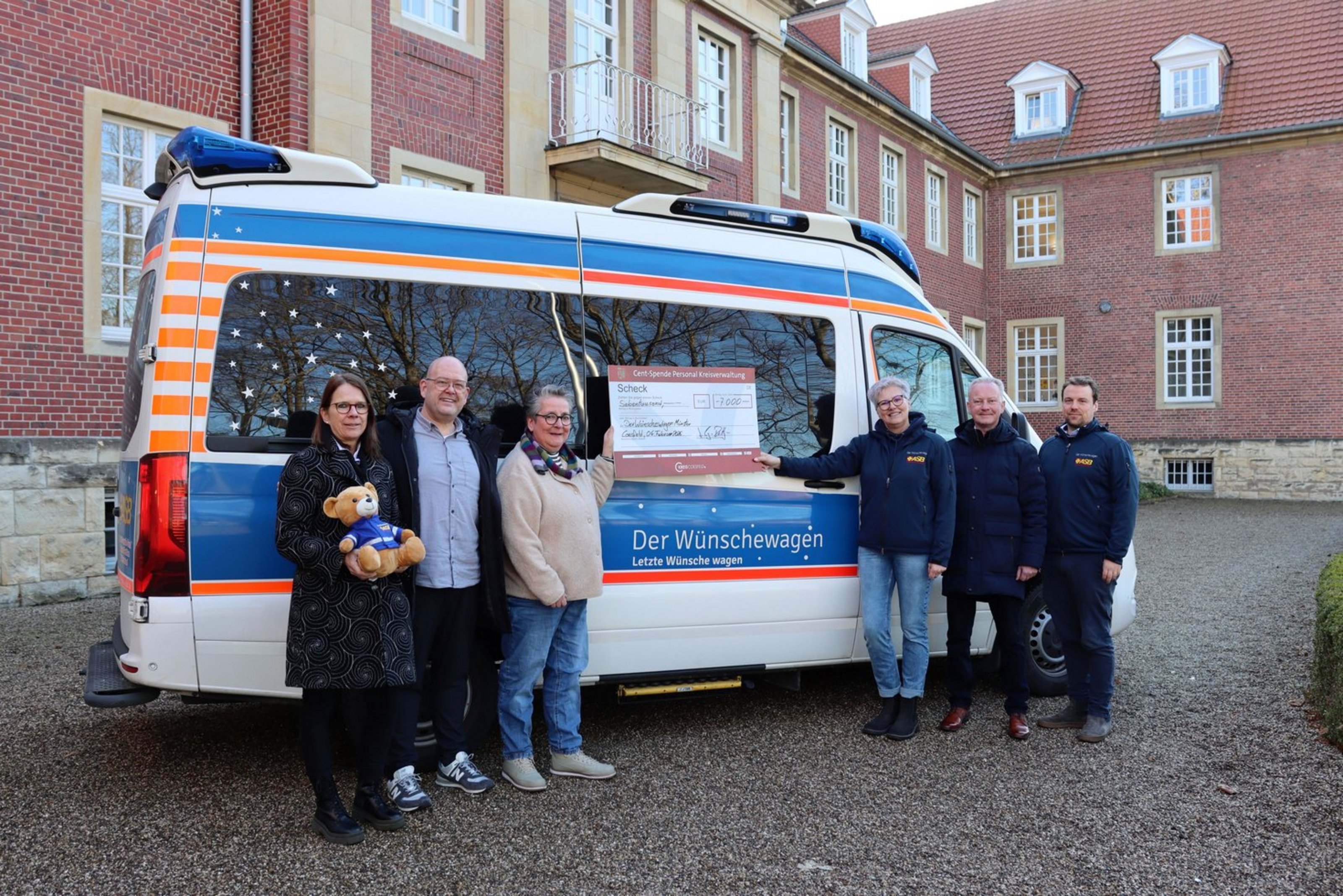 Sechs Personen stehen vor einem Kastenwagen mit der Aufschrift "Der Wünschewagen - Letzte Wünsche wagen". Zwei Personen halten eine Scheck-Tafel. 