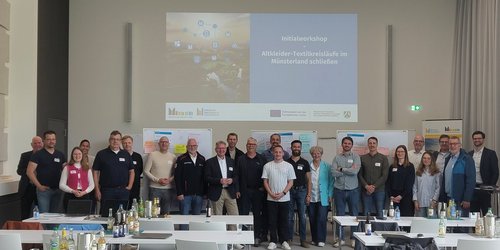 Eine große Gruppe Männer und Frauen steht in einem weißen Raum. Hinter ihnen wird von einem Beamer eine Präsentation an die Wand gestrahlt.