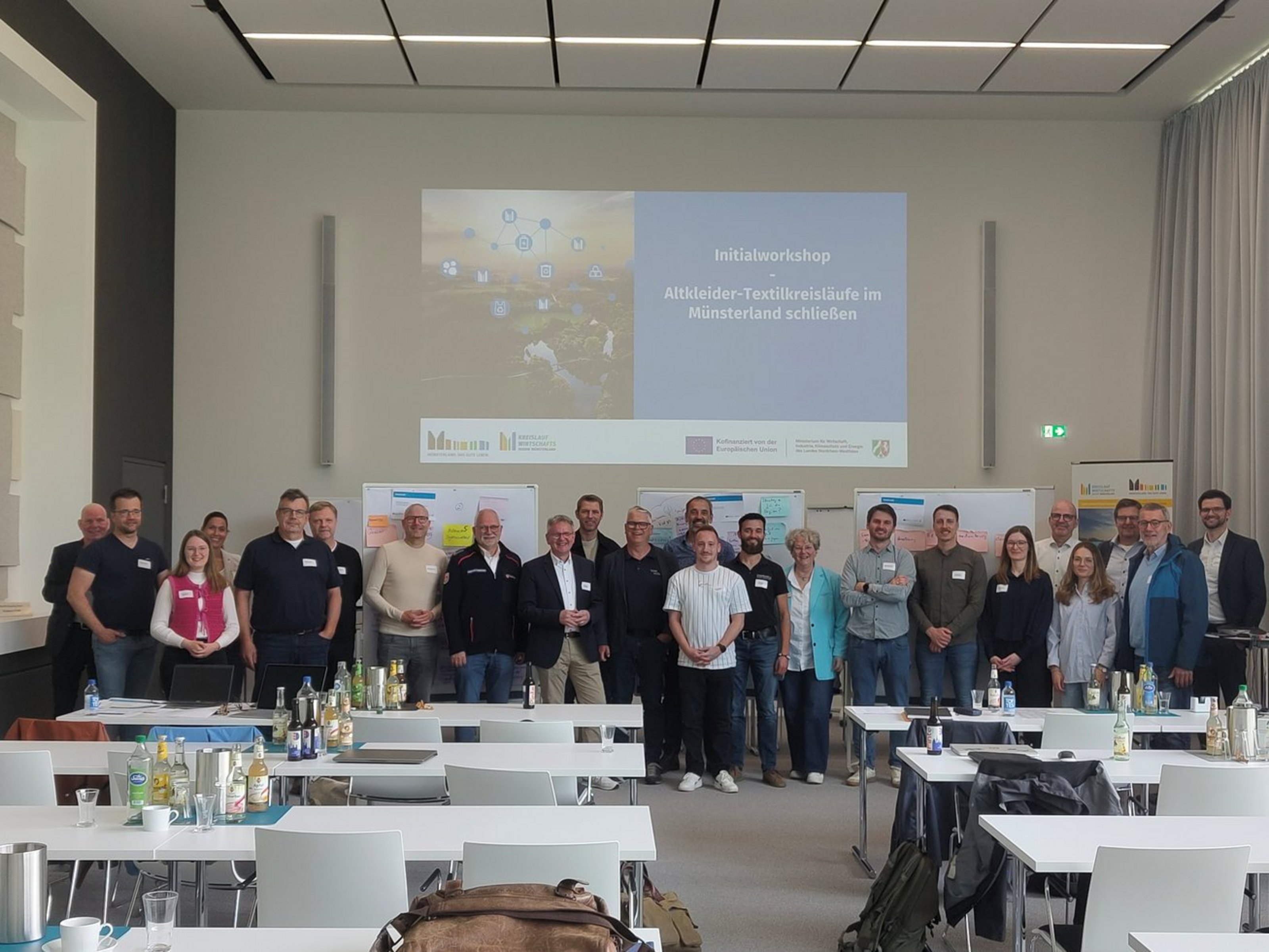 Eine große Gruppe Männer und Frauen steht in einem weißen Raum. Hinter ihnen wird von einem Beamer eine Präsentation an die Wand gestrahlt.