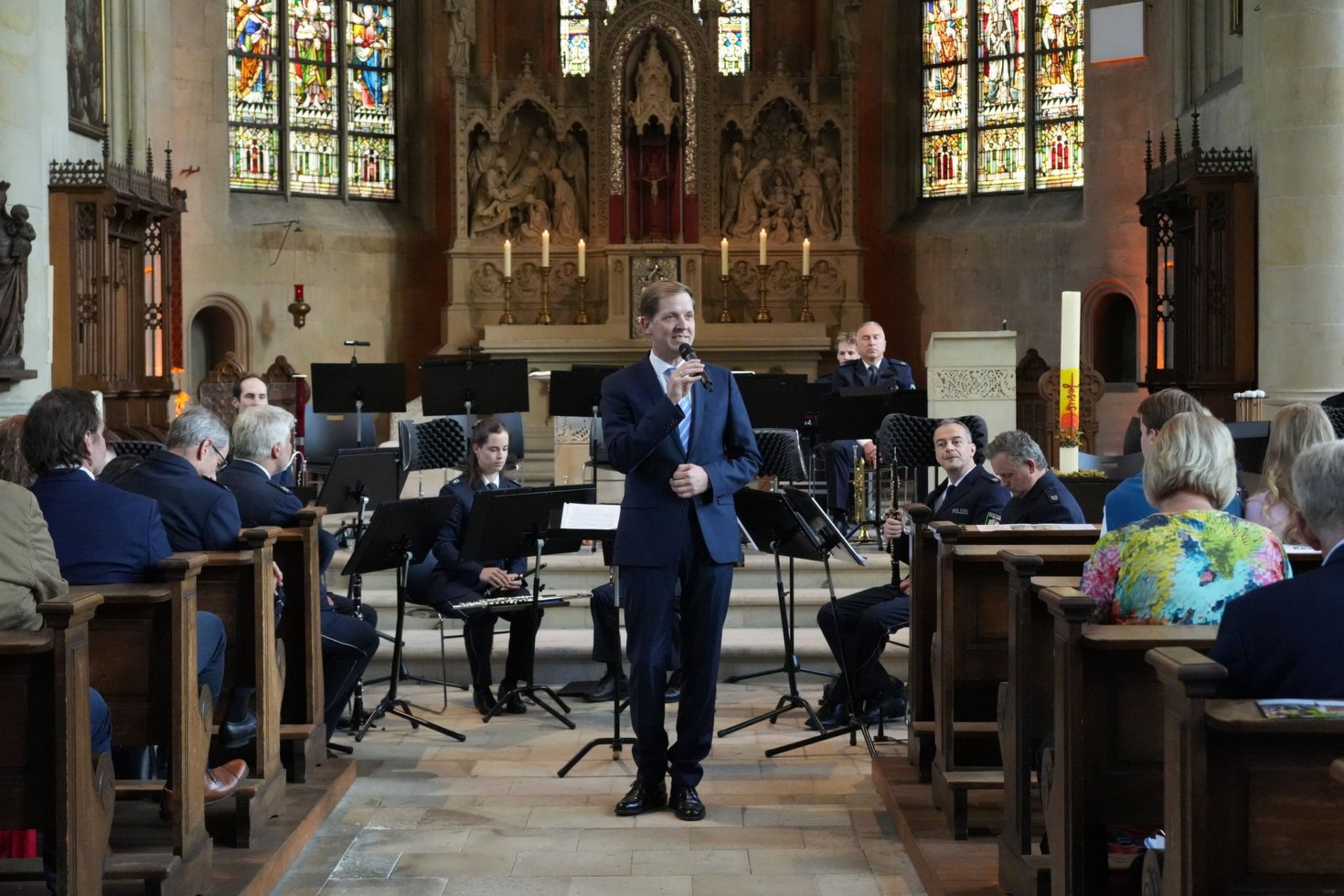 Ein Mann in Anzug steht im Mittelgang einer Kirche mit einem Mikrofon in der Hand. Hinter ihm sitzt ein Orchester. Auf den Kirchenbänken sitzen Menschen.
