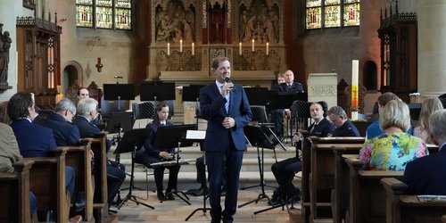 Ein Mann in Anzug steht im Mittelgang einer Kirche mit einem Mikrofon in der Hand. Hinter ihm sitzt ein Orchester. Auf den Kirchenbänken sitzen Menschen.