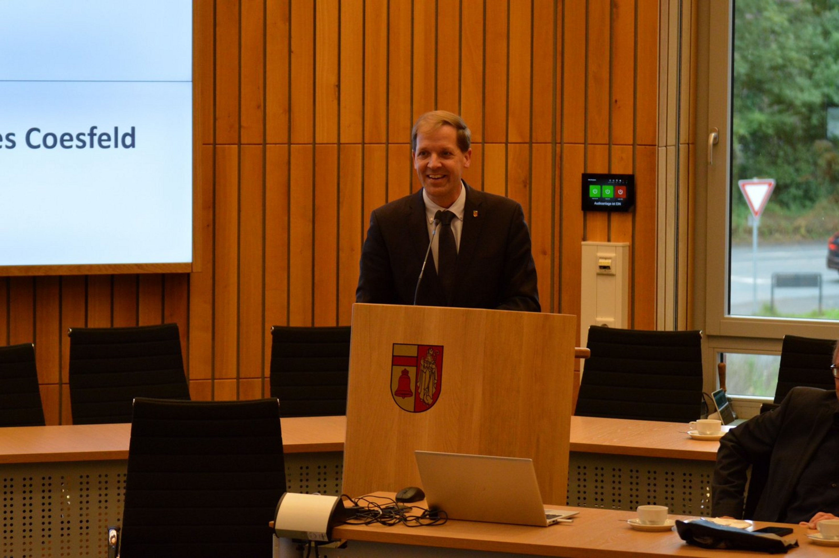 Landrat Dr. Christian Schulze Pellengahr hieß die Trägerinnen und Träger des Bundesverdienstordens im Großen Sitzungssaal des Kreishauses herzlich willkommen (Foto: Kreis Coesfeld) Ein Mann in schwarzem Anzug und mit Krawatte steht hinter einem Rednerpult in einem Raum mit Tischreihen und Stühlen. Die Wand ist mit Holz vertäfelt.