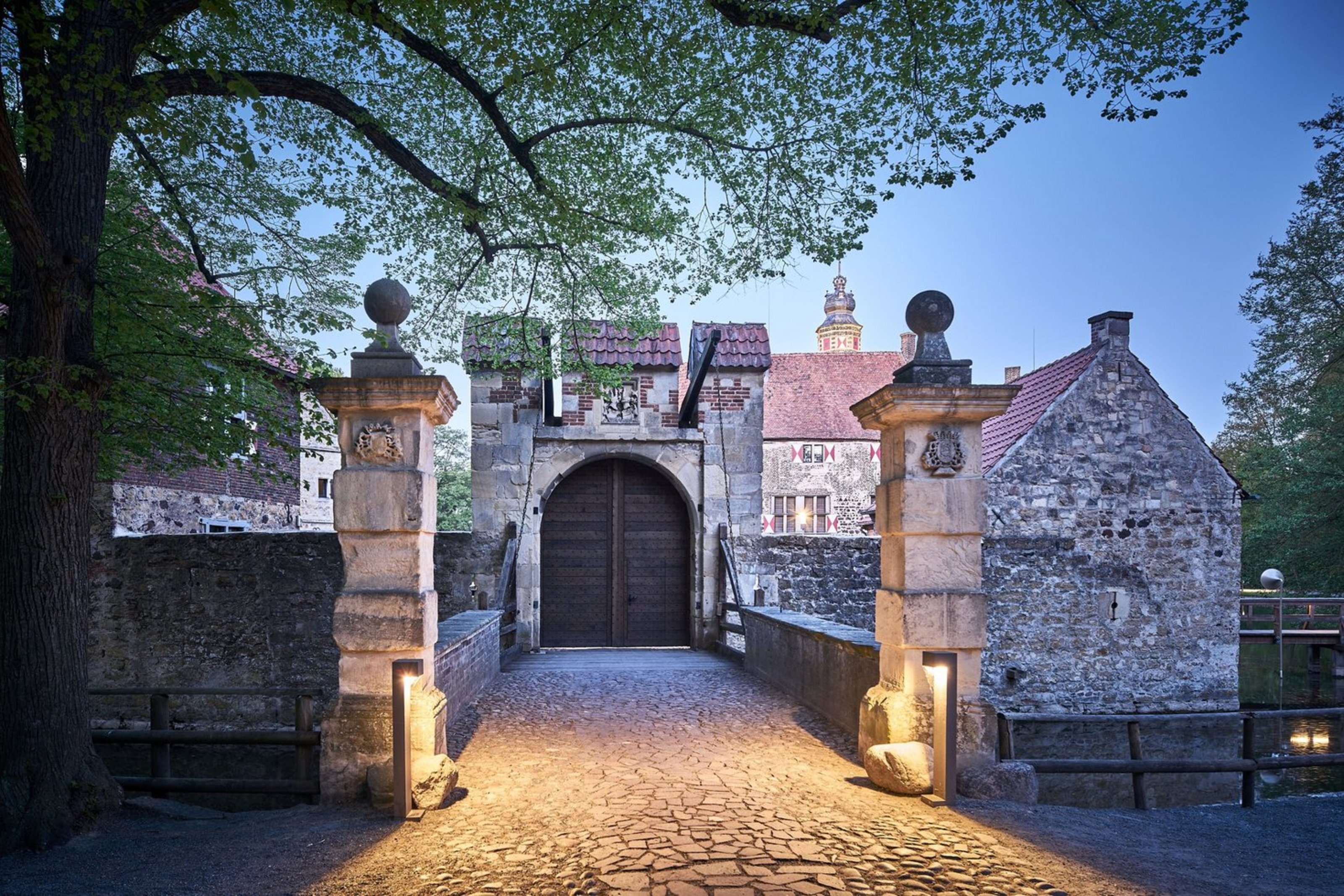 Abendstimmung an der Burg Vischering (Foto: Münsterland e. V. / Philipp Fölting) Ein geschlossenes Burgtor in der Dämmerung wird beleuchtet. Links ragt ein Baum ins Bild.