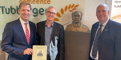 Drei Männer stehen in einer Reihe mit einer Büste. Sie tragen Anzüge. Der linke Mann hält ein Buch in der Hand. Auf der Wand hinter ihnen ist ein Schriftzug, der von den Köpfen verdeckt wird.