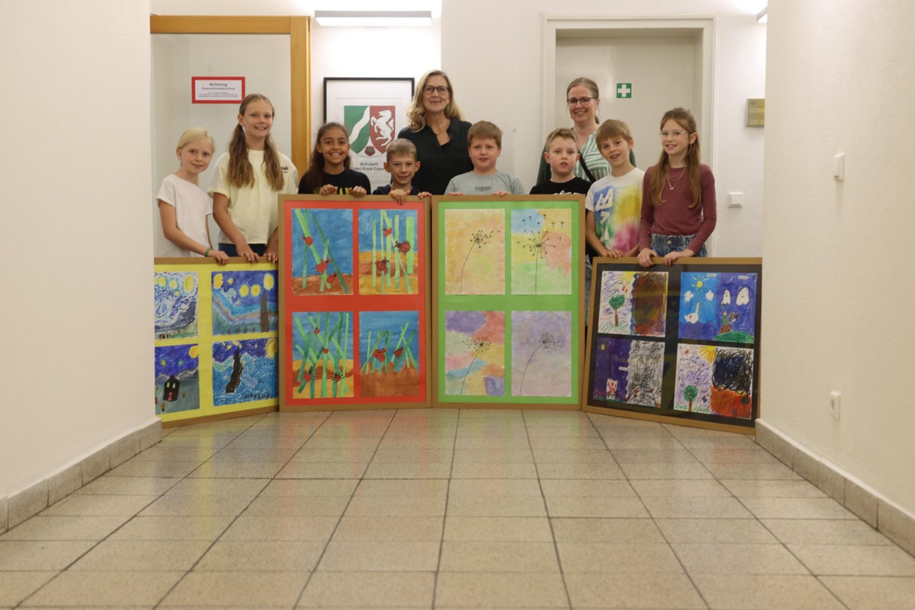Acht Kinder überreichen im Flur des Schulamts vier große, bunte Bilderrahmen mit Kunstwerken. Zwei Erwachsene stehen hinter ihnen. Alle lächeln in die Kamera.
