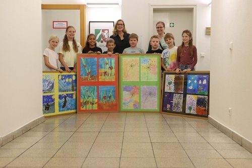Acht Kinder überreichen im Flur des Schulamts vier große, bunte Bilderrahmen mit Kunstwerken. Zwei Erwachsene stehen hinter ihnen. Alle lächeln in die Kamera.