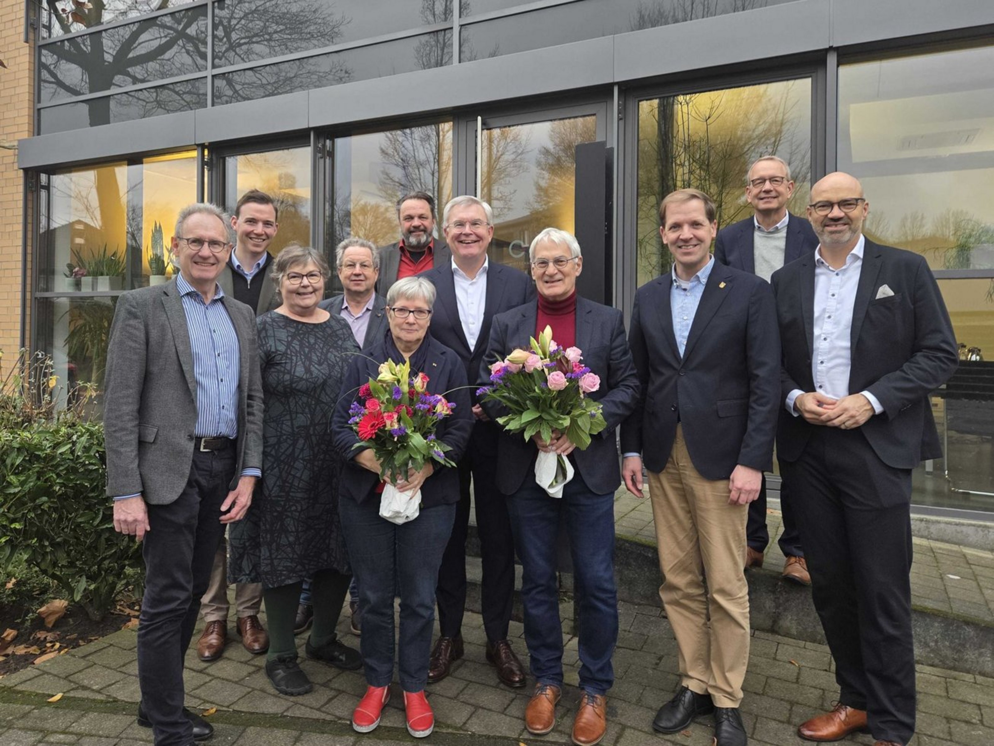 Elf Personen stehen in zwei bis drei Reihen hintereinander. Eine Frau und ein Mann halten jeweils einen Blumenstrauß in der Hand. Hinter der Gruppe befindet sich eine Fensterfront eines Gebäudes. 
