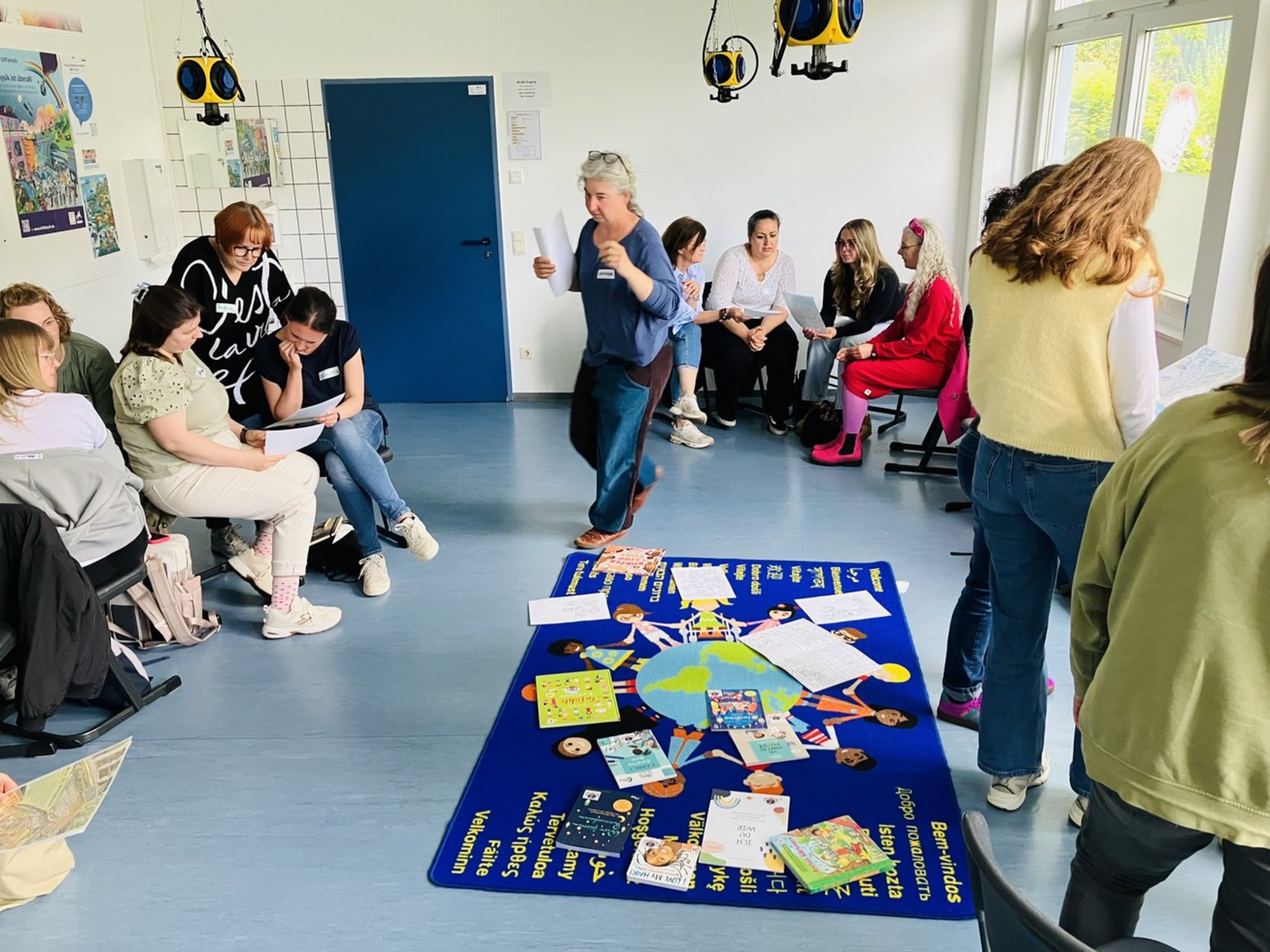 Dreizehn Frauen in einem Klassenraum, in einem Stuhlkreis, sind zu sehen. Sie stehen oder sitzen in Gruppen zusammen und halten Papier in der Hand. Eine Frau läuft von einer Gruppe zur anderen. In der Mitte des Raumes und des Stuhlkreises liegt eine Decke, auf der eine Weltkugel und sich ander Hand haltenden Kinder zu sehen sind. Links und rechts steht in unterschiedlichen Sprachen das Wort "Willkommen".