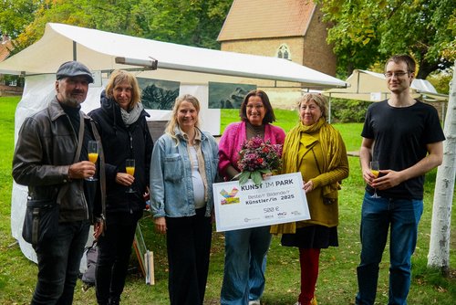 Sechs Personen stehen auf einer grünen Wiese vor einem weißen Zelt. Sie halten Gläser und ein Schild mit der Aufschrift "Kunst im Park. Beste Bildende Künstlerin 2025" in der Hand.