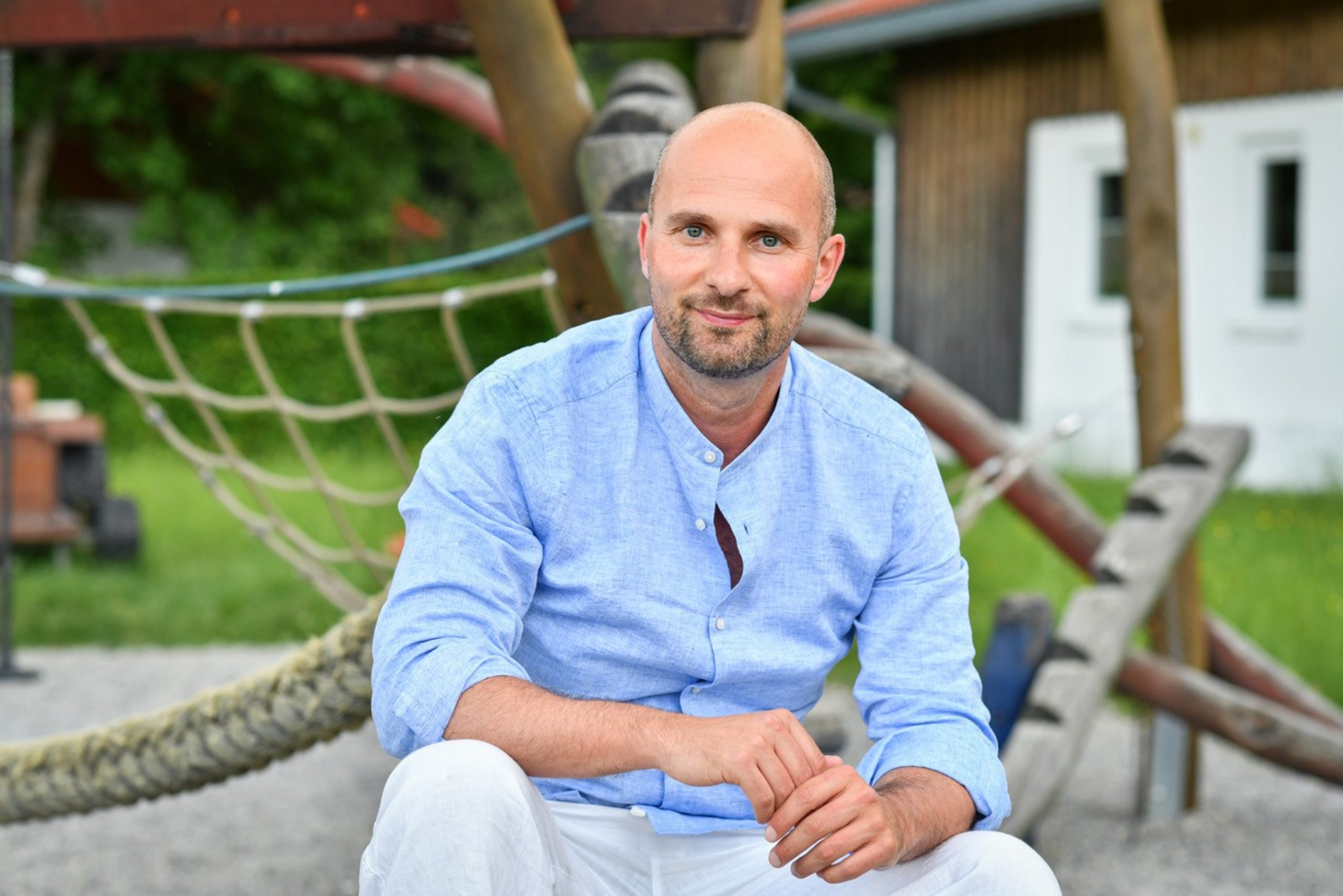 Ein Mann mit blauem Hemd und weißer Hose sitzt entspannt auf einem Spielplatz.