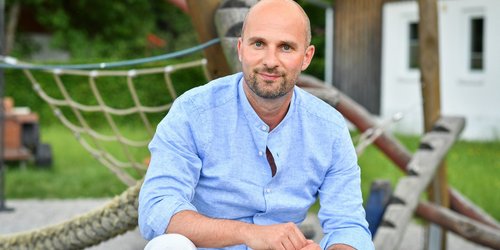 Ein Mann mit blauem Hemd und weißer Hose sitzt entspannt auf einem Spielplatz.