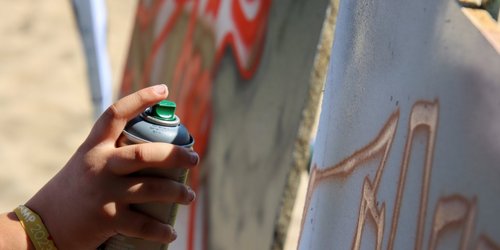 Eine Kinderhand hält eine Spraydose und sprüht Farbe auf eine Leinwand.