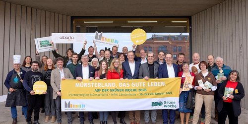 Eine Gruppe von vielen Personen steht in drei Reihen vor einem großen Fenster eines Gebäudes. Die Personen in der ersten Reihe halten einen Banner mit der Aufschrift "Münsterland. Das gute Leben. Auf der Grünen Woche 2026. Partnerregion Münsterland. MRW Länderhalle. 16. bis 25. Januar 2026."