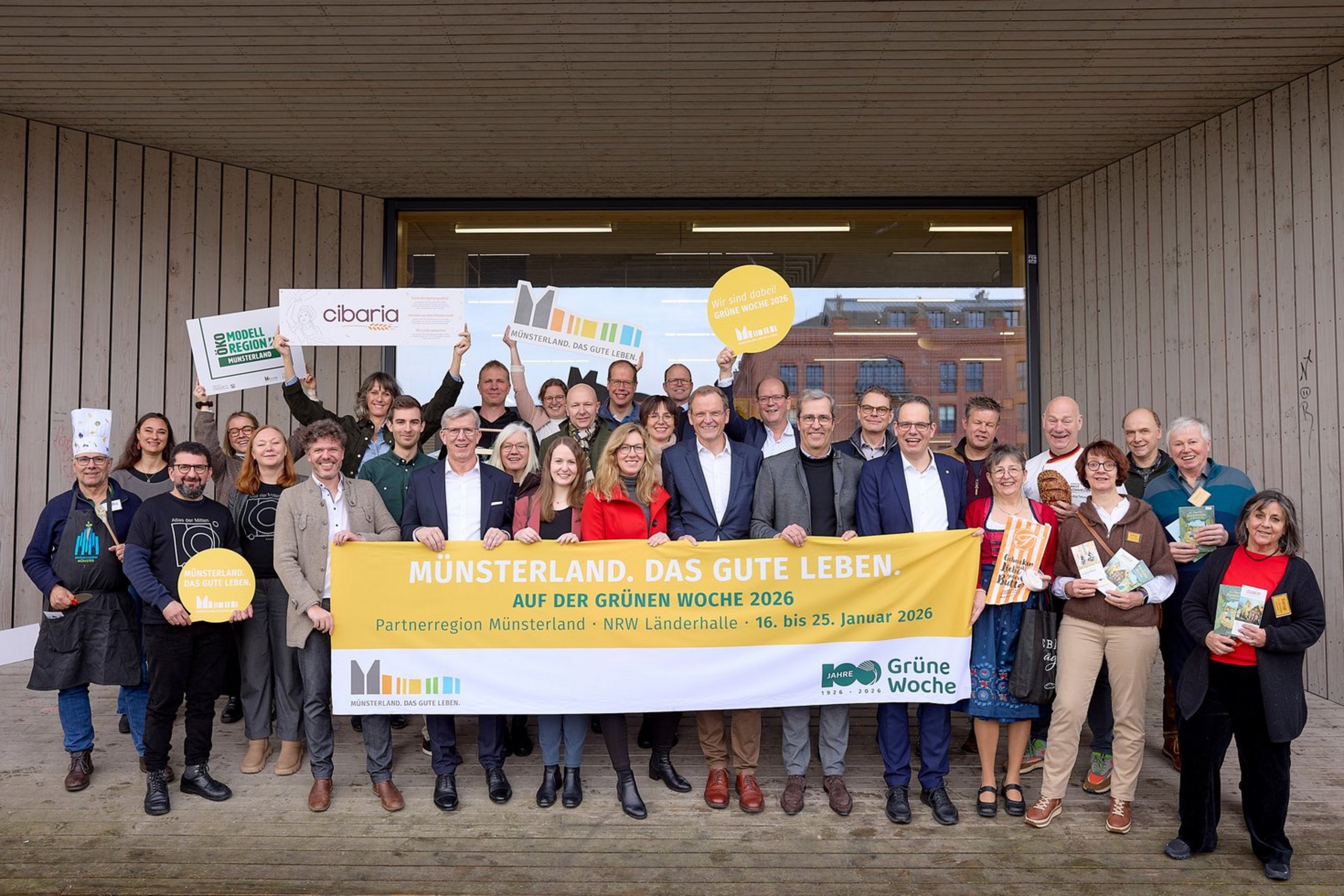 Eine Gruppe von vielen Personen steht in drei Reihen vor einem großen Fenster eines Gebäudes. Die Personen in der ersten Reihe halten einen Banner mit der Aufschrift "Münsterland. Das gute Leben. Auf der Grünen Woche 2026. Partnerregion Münsterland. MRW Länderhalle. 16. bis 25. Januar 2026."
