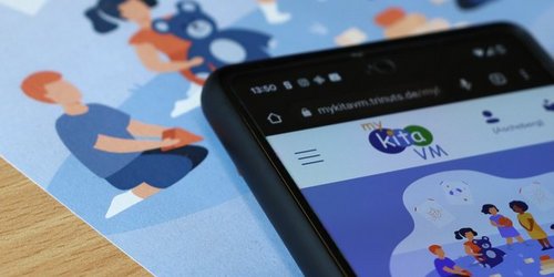 Man sieht einen blauen Flyer auf dem ein Smartphone liegt. Auf dem Handy ist die App mykita vm geöffnet.