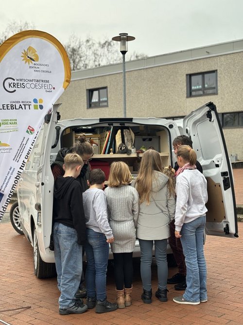 Eine Gruppe von Kindern steht mit einer Frau vor geöffneten Hecktüren eines weißen Autos. Sie alle schauen in den Kofferraum.