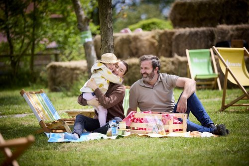 Eine Frau, ein Mann und ein Kind sitzen auf einer Picknickdecke auf einer Grünfläche. Im Hintergrund sind bunte Liegestühle, Bäume und eine Mauer aus Strohballen zu sehen. Das Kind und die Frau umarmen sich. Der Mann schaut lächelnd zu den beiden. Auf der Decke stehen drei kleine Gläser, eine Wasserflasche und eine Holzkiste mit einem rot-weiß kariertem Tuch.